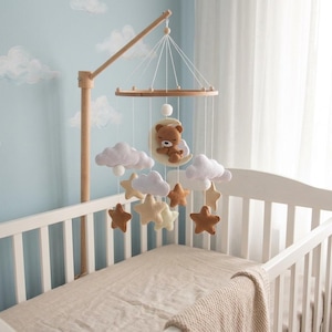 May include: A baby crib mobile with a wooden frame and hanging felt decorations. The mobile features a sleeping teddy bear on a crescent moon, white clouds, and stars in shades of brown and yellow. The crib is white with a beige blanket.