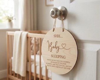 May include: A round wooden sign with the words "SHH... Baby SLEEPING" engraved on it, hanging from a door handle. The sign also includes a request to not ring the doorbell or knock. The sign is hanging on a white door.