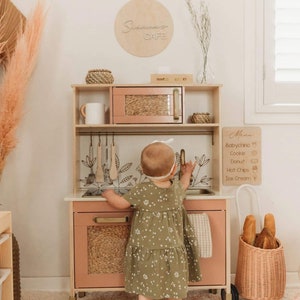 Puede incluir: Un ni&ntilde;o juega con una cocina de juguete de madera. La cocina est&aacute; pintada en tonos rosa suave y madera natural, con microondas, fregadero y elementos decorativos. Un letrero encima de la cocina dice "Sienna's Cafe".