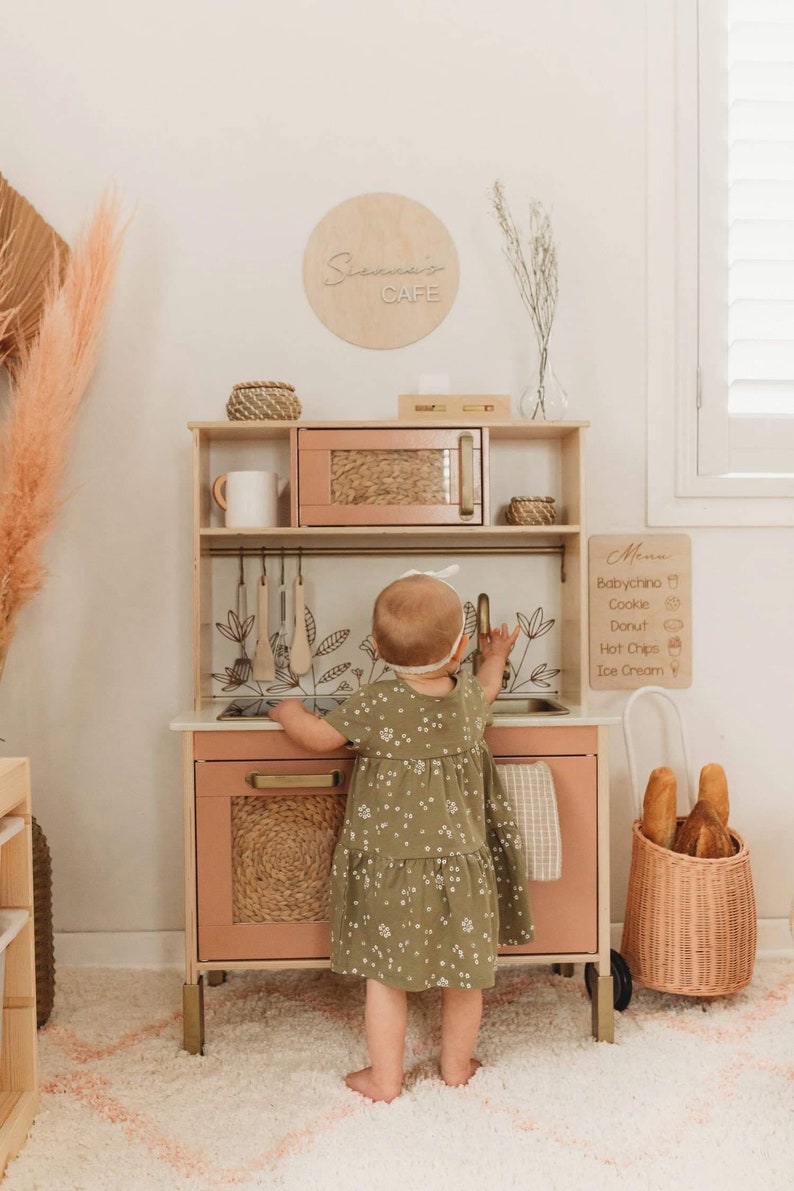 Puede incluir: Un ni&ntilde;o juega con una cocina de juguete de madera. La cocina est&aacute; pintada en tonos rosa suave y madera natural, con microondas, fregadero y elementos decorativos. Un letrero encima de la cocina dice "Sienna's Cafe".