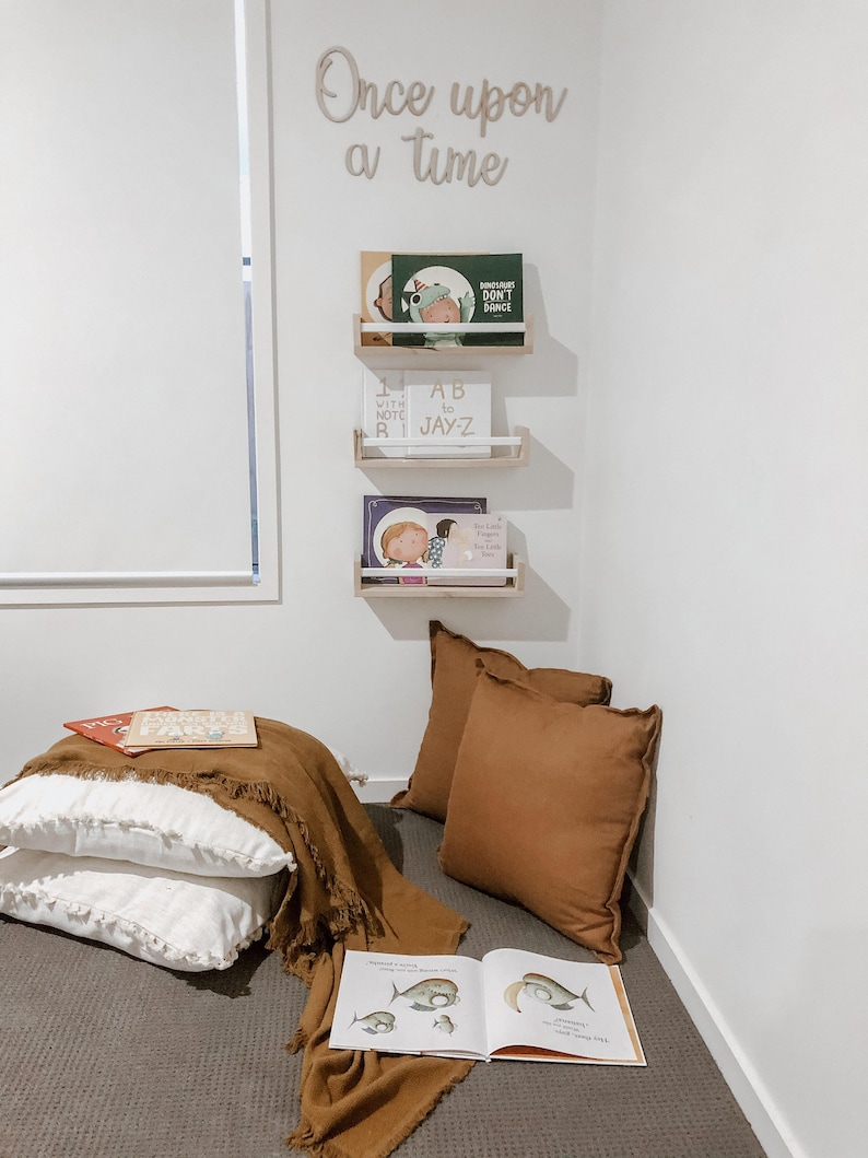 May include: A cozy reading nook with a brown rug, two white pillows with fringe, a brown throw blanket, and three bookshelves with children's books. The shelves are mounted on a white wall with the words "Once upon a time" written in cursive.