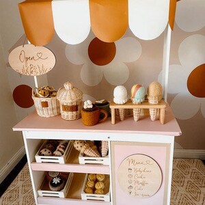 May include: A white wooden play stand with a brown and white awning, shelves, and a sign that says "Open". The stand is decorated with a brown and white daisy pattern. There are various play food items on the shelves, including pastries, cookies, and ice cream cones.