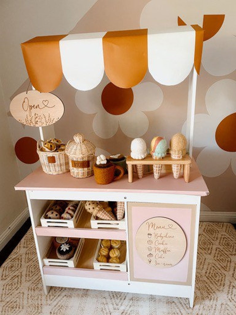 May include: A white wooden play stand with a brown and white awning, shelves, and a sign that says "Open". The stand is decorated with a brown and white daisy pattern. There are various play food items on the shelves, including pastries, cookies, and ice cream cones.