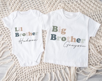 Puede incluir: Dos camisetas blancas con el texto "Lil Brother Hudson" y "Big Brother Grayson" en letras de colores. Las camisetas están sobre una superficie de crochet beige.