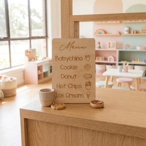 Puede incluir: Un tablero de men&uacute; de juego de madera con texto grabado e iconos de alimentos, que incluyen un babyccino, galleta, donut, patatas fritas y helado. Una peque&ntilde;a taza de madera, galleta y donut est&aacute;n en el mostrador. El men&uacute; es parte de un juego.