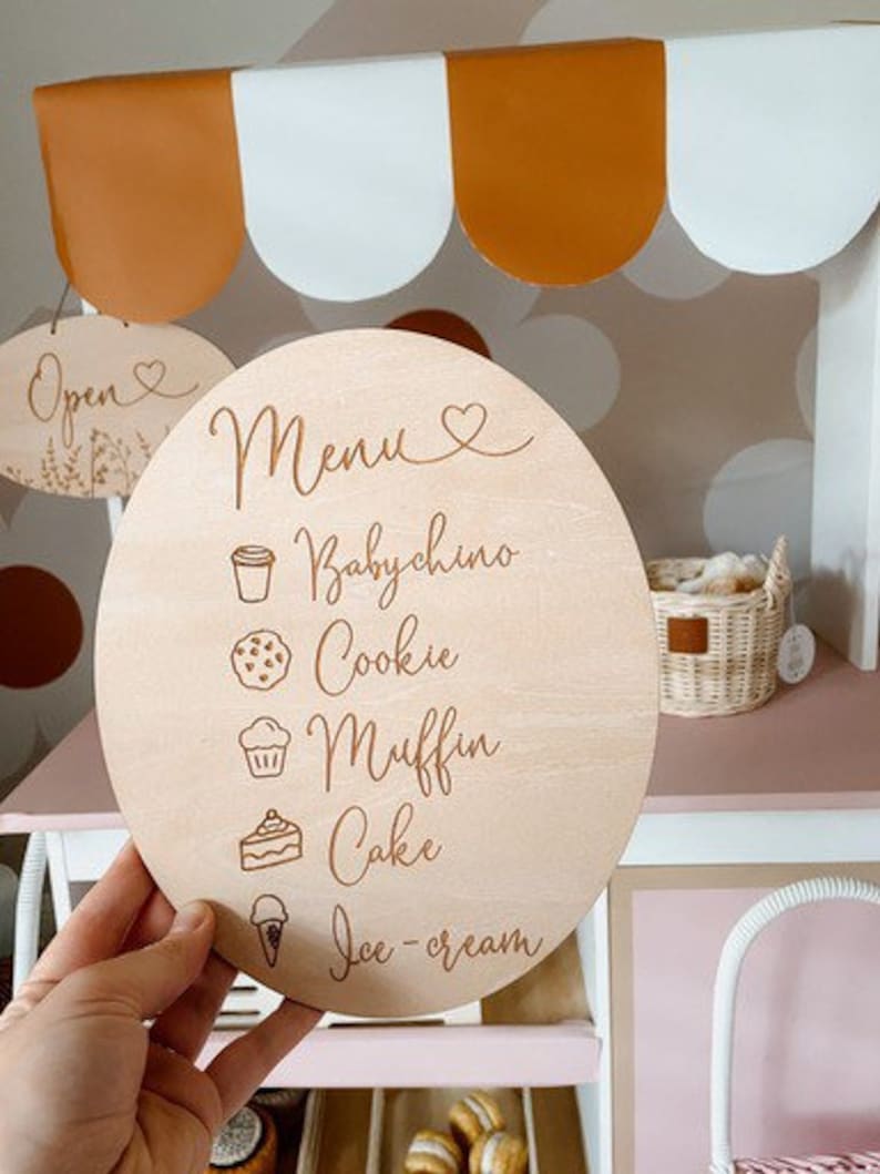 May include: A round wooden sign with a menu for a pretend play shop. The menu includes: Babychino, Cookie, Muffin, Cake, and Ice-cream.