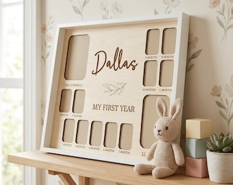 Puede incluir: Un marco de fotos de bebé de madera con marco blanco con el nombre "Dallas" y el texto "My First Year". Tiene espacios para fotos desde "Hello World" hasta los 12 meses. Un conejito de peluche y bloques decorativos están cerca.