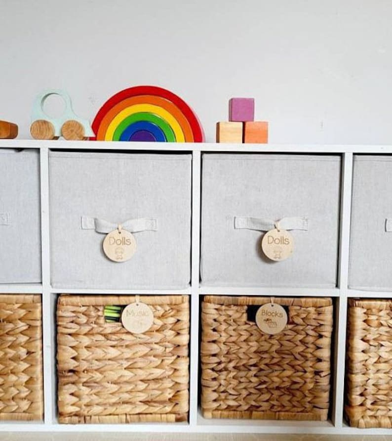 May include: White cube storage shelves with woven baskets labeled "Dolls", "Blocks", and "Music". The shelves are decorated with a rainbow arch, a toy car, and wooden blocks.