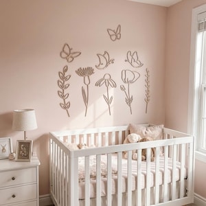 May include: A nursery scene featuring a white cot, a white chest of drawers, and wall decorations. The wall has wooden cutouts of flowers and butterflies. The cot contains a pink blanket and soft toys. A lamp and framed photos are on the chest.