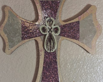 May include: A decorative wooden cross with a distressed finish. The cross is painted in shades of beige and pink with glitter accents. A small white flower is attached to the center of the cross.