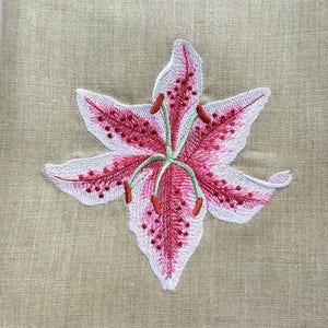 Pu&ograve; includere: Un asciugamano da cucina in lino beige con un ricamo di giglio rosa e bianco.