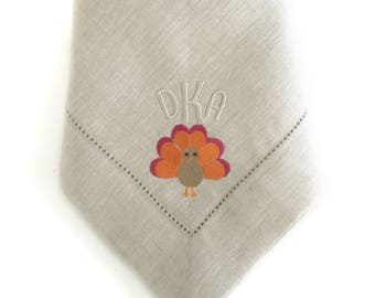 May include: A beige linen napkin with a turkey and the letters "DKA" embroidered in orange, brown, and pink.