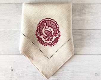 May include: A folded beige linen napkin with a decorative embroidered turkey design in burgundy. The turkey is surrounded by a wreath, and the napkin has a stitched border. Ideal for Thanksgiving or fall decor.