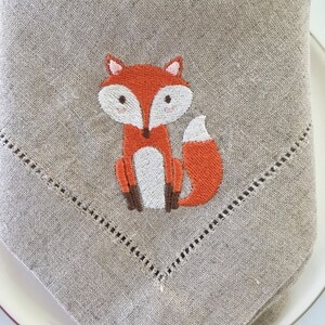 May include: A beige linen napkin featuring an embroidered orange fox design. The fox has a white belly and brown legs.