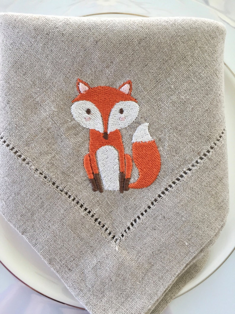 May include: A beige linen napkin featuring an embroidered orange fox design. The fox has a white belly and brown legs.