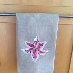 Pu&ograve; includere: Un asciugamano da cucina in lino beige con un giglio rosa ricamato al centro.