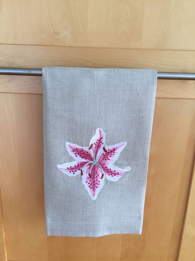 Pu&ograve; includere: Un asciugamano da cucina in lino beige con un giglio rosa ricamato al centro.