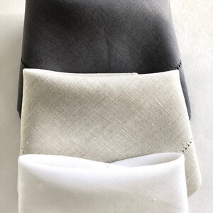 May include: Three linen napkins folded in a triangle shape. The top napkin is dark gray, the middle napkin is light gray, and the bottom napkin is white.