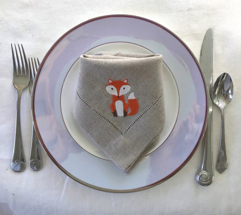 May include: A white dinner plate with a light purple rim, a linen napkin with an embroidered orange fox, and silver cutlery.