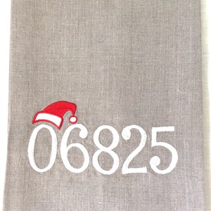 Puede incluir: Una toalla de cocina de lino gris con un número 06825 bordado en blanco y un gorro de Papá Noel rojo.