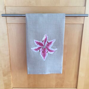 Pu&ograve; includere: Un asciugamano da cucina in lino beige con un giglio rosa ricamato.