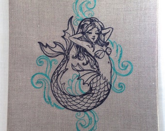 May include: A linen tea towel with an embroidered design of a mermaid with teal blue waves and swirls.