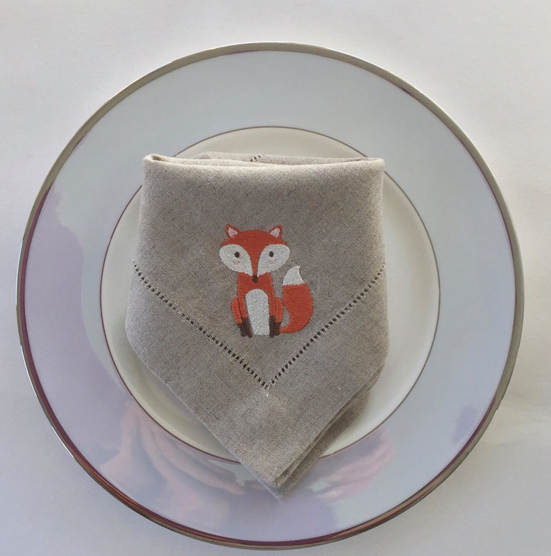 May include: A beige linen napkin with an embroidered orange fox design. The napkin is folded and placed on a white plate with a silver rim.