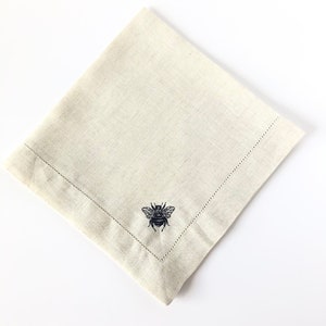 May include: A beige linen napkin with a black embroidered bee design.