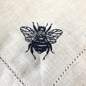 May include: A white linen napkin with a black embroidered bee design. The napkin has a delicate lace trim around the edges.