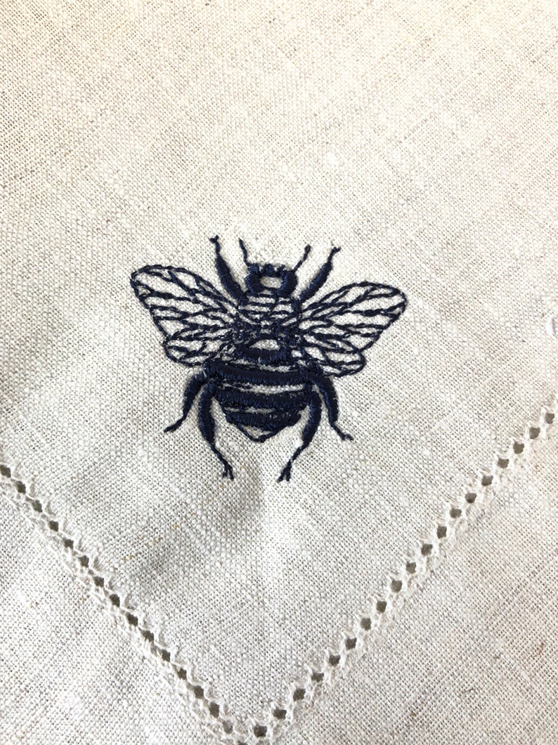 May include: A white linen napkin with a black embroidered bee design. The napkin has a delicate lace trim around the edges.