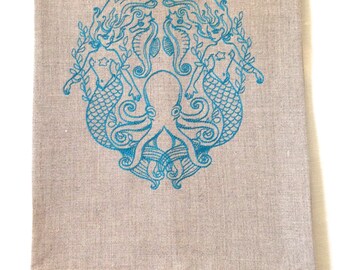 May include: A teal blue linen tea towel with an embroidered design of two mermaids flanking an octopus. The mermaids have flowing hair and tails, and the octopus has eight tentacles.