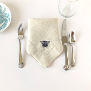 May include: A beige linen napkin with a dark blue embroidered bee design. The napkin is folded into a triangle shape and is placed on a white surface with a fork, knife, and spoon.
