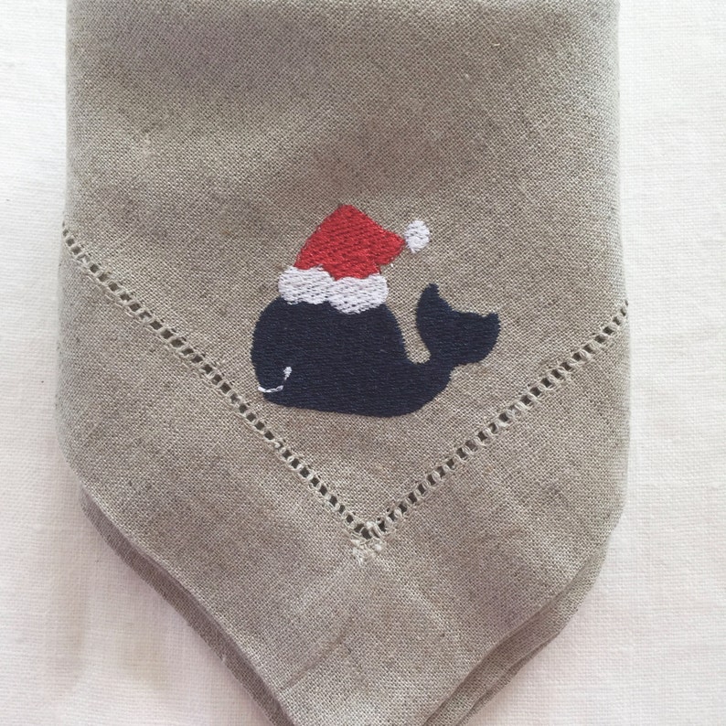 May include: A gray linen napkin with a white stitched border. A blue whale with a red and white Santa hat is embroidered on the napkin.
