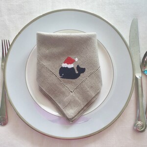 May include: A beige linen napkin with a whale wearing a Santa hat embroidered on it. The napkin is folded and placed on a white plate with a silver rim.