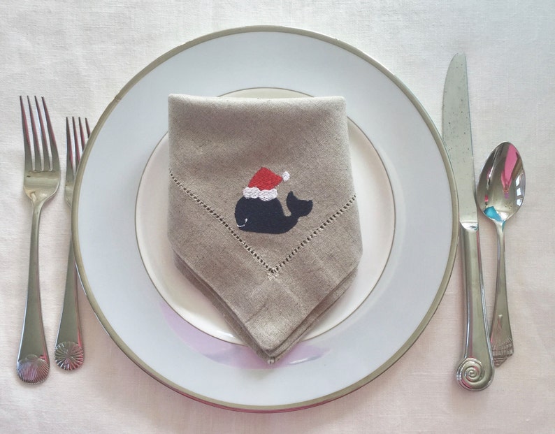 May include: A beige linen napkin with a whale wearing a Santa hat embroidered on it. The napkin is folded and placed on a white plate with a silver rim.