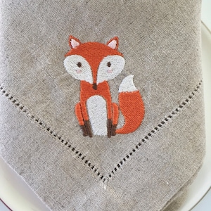 May include: A beige linen napkin featuring an embroidered orange fox design. The fox has a white belly and brown legs.