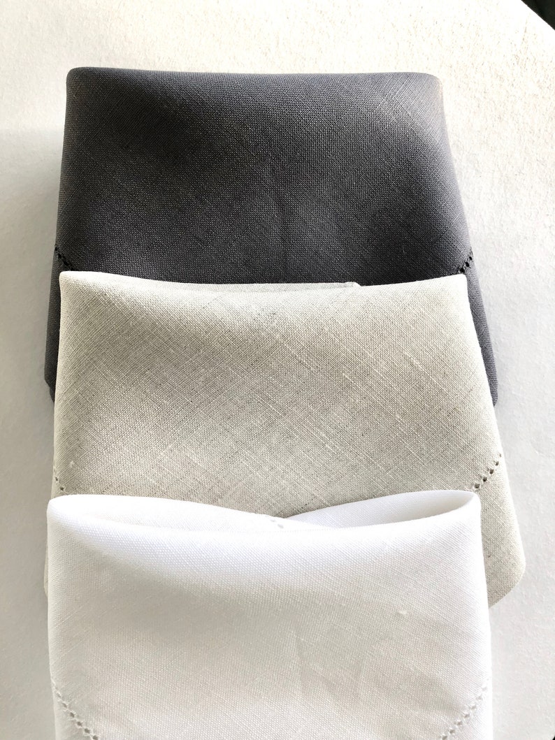 May include: Three linen napkins folded in a triangular shape. The top napkin is dark grey, the middle napkin is light grey, and the bottom napkin is white.