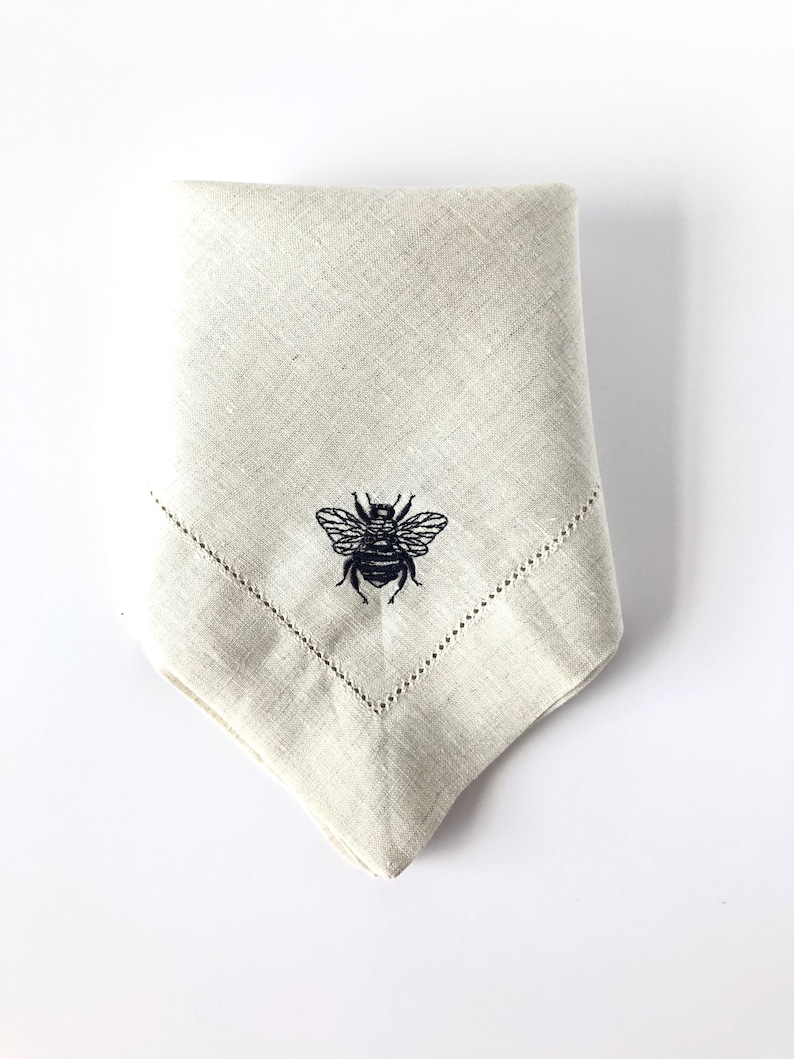 May include: A beige linen napkin with a black embroidered bee design.