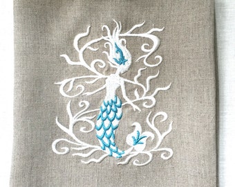 May include: A beige linen tea towel with a white embroidered mermaid design. The mermaid's tail is turquoise blue.