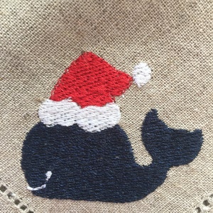 May include: Embroidered whale wearing a red and white Santa hat on a beige fabric background.