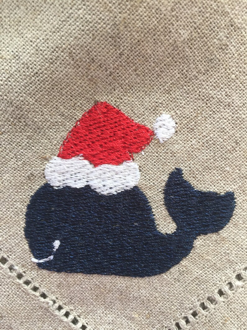 May include: Embroidered whale wearing a red and white Santa hat on a beige fabric background.