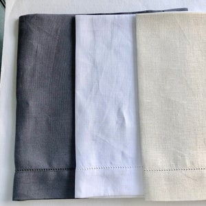 May include: Three linen napkins in different colors: dark gray, white, and light beige. The napkins have a subtle texture and a stitched edge.