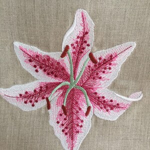 Pu&ograve; includere: Fiore di giglio ricamato rosa e bianco su uno sfondo di tessuto di lino beige.