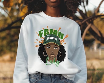May include: White sweatshirt featuring a graphic of a woman wearing a green and gold baseball cap with "FAMU HBCU" on it. The graphic also includes the word "Lady" in green script and orange paint splatters.