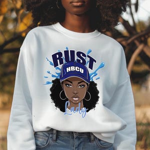 May include: White sweatshirt with a blue and white graphic of a woman wearing a blue hat with the text "HBCU" on it. The text "Rust Lady" is below the graphic.