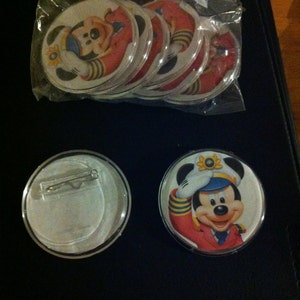May include: A set of eight round, clear plastic buttons with a printed image of Mickey Mouse dressed as a sailor. The buttons have a silver safety pin on the back. One button is shown separately with a close-up of Mickey Mouse.