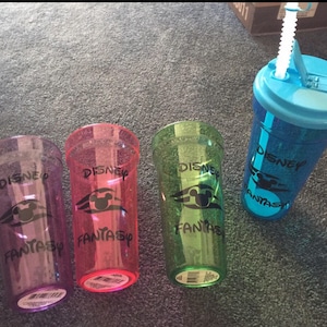 May include: Four clear plastic cups with lids and straws. The cups are decorated with a Disney logo and the words "Disney Fantasy". The cups are in different colors: purple, pink, green, and blue.
