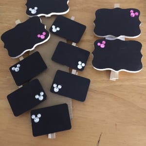 Könnte beinhalten: Ein Set aus neun schwarzen Mini-Kreidetafeln mit Holzklammern. Jede Kreidetafel hat ein Mickey-Mouse-Kopfdesign mit rosa und weißen Strasssteinen.