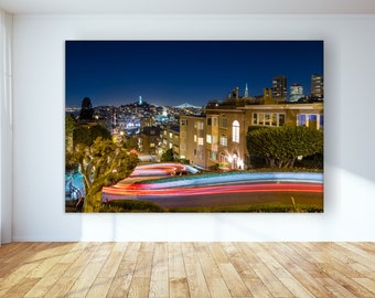May include: A night time view of a winding road in San Francisco with the city skyline in the background. The road is lit by streetlights and the headlights of cars driving up the hill. The Golden Gate Bridge is visible in the distance.
