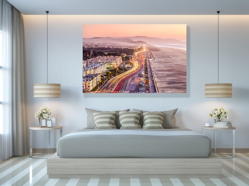 May include: An aerial view of a city at sunset with a highway winding through the city and along the coast. The highway is lit up with car headlights and taillights, creating a long streak of light. The beach is sandy and the ocean is calm.